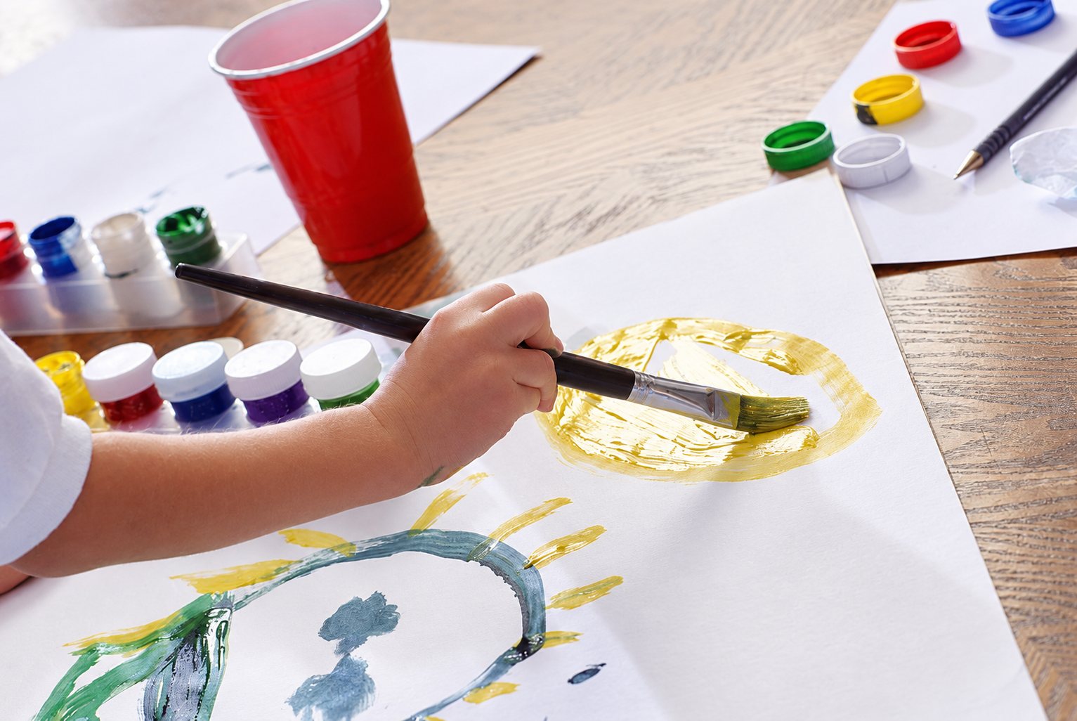 La main d’un enfant qui peint au pinceau sur une grande feuille, entourée de pots de peinture